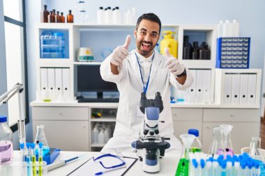 Laboratuvarda çalışan, sakallı, genç İspanyol bir adam. Elleriyle olumlu jest yapıyor. Gülümsüyor ve başarı için seviniyor. kazanan jest. 