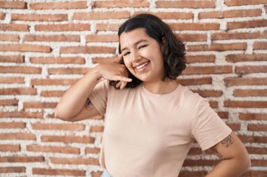 Young hispanic woman standing over bricks wall smiling doing phone gesture with hand and fingers like talking on the telephone. communicating concepts. 