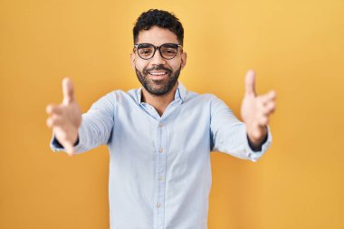 Hispanic man with beard standing over yellow background looking at the camera smiling with open arms for hug. cheerful expression embracing happiness. 
