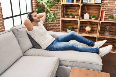 Genç İspanyol adam ellerini başının üstüne koyup evde müzik dinliyor.