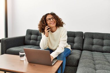 Orta yaşlı İspanyol kadın evde akıllı telefondan dizüstü bilgisayar kullanıyor.