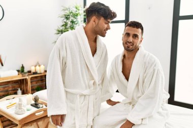 Two hispanic men couple wearing bathrobe sitting on massage table at beauty center