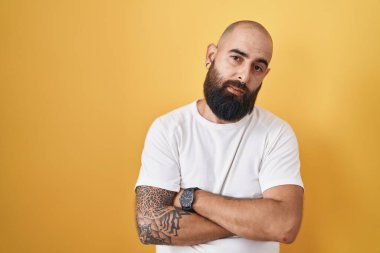 Young hispanic man with beard and tattoos standing over yellow background looking sleepy and tired, exhausted for fatigue and hangover, lazy eyes in the morning. 