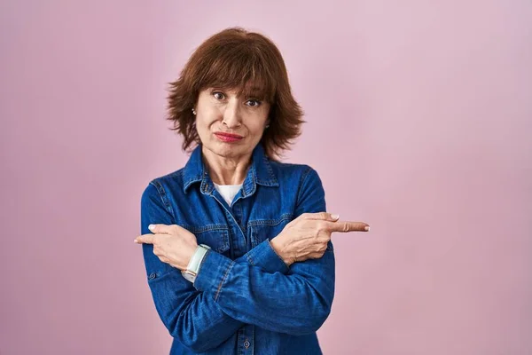 Middle age woman standing over pink background pointing to both sides with fingers, different direction disagree