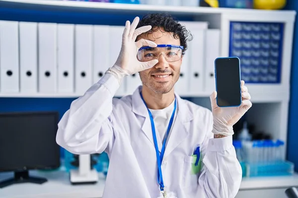 ヒスパニック系の男仕事で科学者の研究室を示していますスマートフォンの画面笑顔幸せな行いますOk Signとともに手で目を通して指 — ストック写真