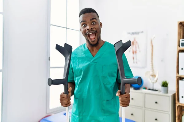 Young African Physiotherapist Man Holding Crutches Celebrating Crazy Amazed Success — Stock Photo, Image