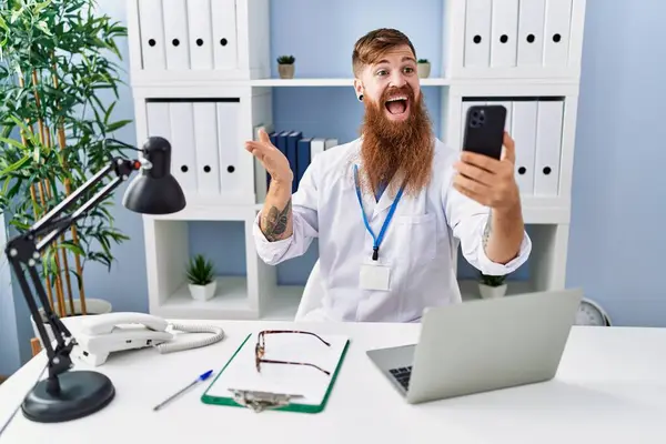 ビデオ通話を行う長い髭を持つ赤毛の男は幸せな笑顔で達成を祝うスマートフォンと相談し 手を上げと勝者の式 — ストック写真