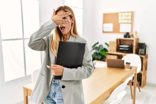 Blonde business woman at the office peeking in shock covering face and eyes with hand, looking through fingers with embarrassed expression.