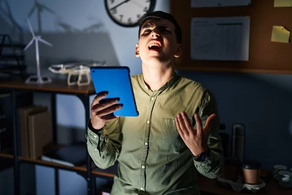 Non binary person using touchpad device at night crazy and mad shouting and yelling with aggressive expression and arms raised. frustration concept.