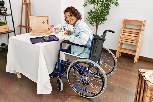 Middle age hispanic disabled artist woman drawing sitting on wheelchair at art studio.