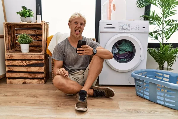 Young Blond Man Doing Laundry Using Smartphone Sticking Tongue Out — Stok fotoğraf