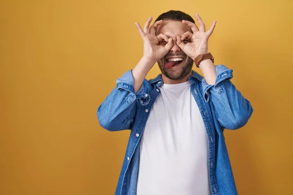 Hispanic Man Standing Yellow Background Doing Gesture Binoculars Sticking Tongue — Stock fotografie