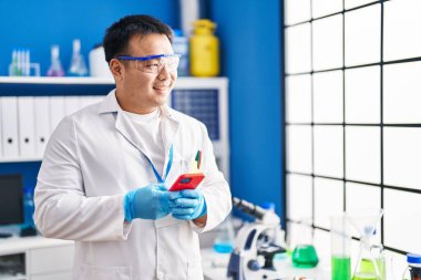 Young chinese man wearing scientist uniform using smartphone at laboratory