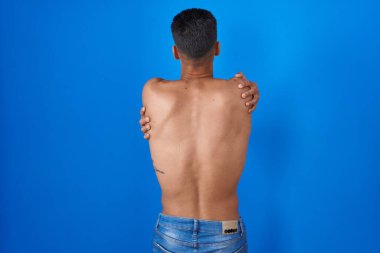 Mavi arka planda tişörtsüz duran İspanyol genç adam kendine sarılıyor mutlu ve pozitiftir. Sevgi ve özgüven. 