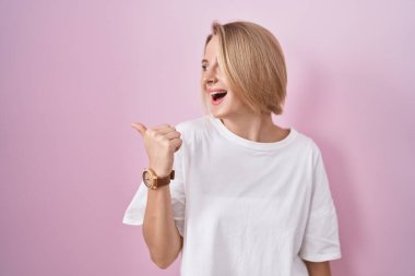 Pembe arka planda duran, gülümseyen, gülümseyen ve baş parmağıyla yan tarafı gösteren beyaz bir kadın.. 