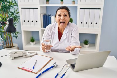 Hispanic mature doctor woman holding pill and glass of water celebrating crazy and amazed for success with open eyes screaming excited. 