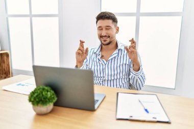 Ofiste çalışan sakallı yakışıklı genç adam diz üstü bilgisayar kullanarak umut ve gözlerin kapalı bir şekilde gülümseyerek işaret ediyor. Şans ve batıl inançlar. 