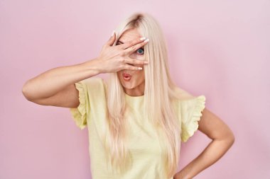 Caucasian woman standing over pink background peeking in shock covering face and eyes with hand, looking through fingers with embarrassed expression. 