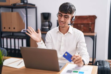 Young hispanic man working using computer laptop holding credit card smiling cheerful presenting and pointing with palm of hand looking at the camera. 