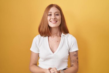 Young redhead woman standing over yellow background with hands together and crossed fingers smiling relaxed and cheerful. success and optimistic 