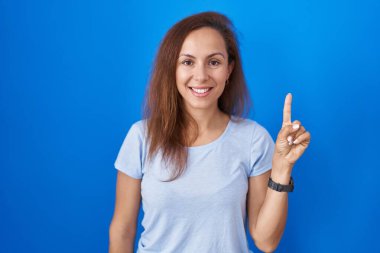 Brunette woman standing over blue background showing and pointing up with finger number one while smiling confident and happy. 