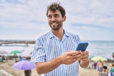 Genç İspanyol adam sahilde akıllı telefon kullanarak mutlu bir şekilde gülümsüyor.