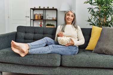 Orta yaşlı, kır saçlı, gülümseyen, kendine güvenen, evde film izleyen bir kadın.