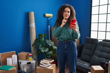 Yeni evdeki Latin kökenli genç kadın akıllı telefon kullanıyor ciddi bir yüz ifadesi, eli çenesinde soru soran, kafa karıştırıcı bir fikir hakkında düşünceli. 