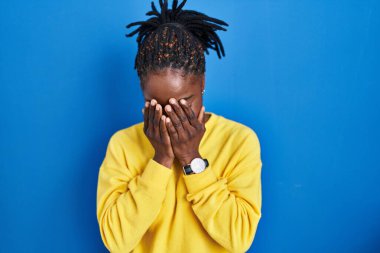Beautiful black woman standing over blue background with sad expression covering face with hands while crying. depression concept. 