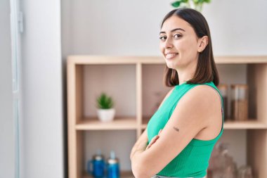 Genç İspanyol kadın gülümsüyor. Kollarını kavuşturup evde duruyor.