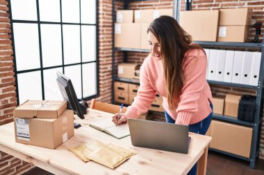 Young beautiful hispanic woman ecommerce business worker using laptop writing on notebook at office