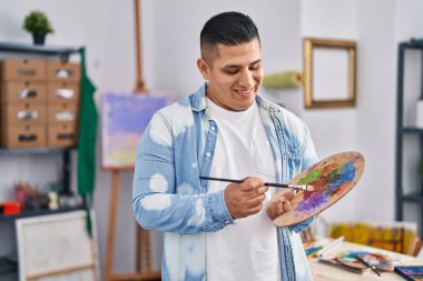 Sanat stüdyosunda palet ve boya fırçasıyla gülümseyen genç Latin sanatçı.