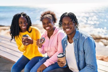 African american friends having breakfast sitting on bench at seaside