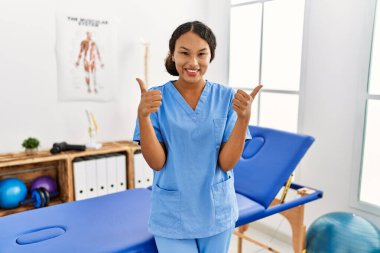 Güzel İspanyol fizyoterapist kadın. Ağrı tedavisi kliniğindeki başarı işareti. Elleriyle olumlu jest yapıyor. Neşeli ifade ve kazanan jest. 