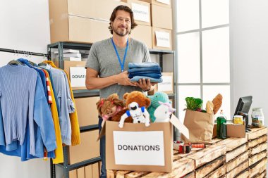 Orta yaşlı beyaz bir adam yardım merkezinde katlanmış giysiler taşıyarak gülümsüyor.