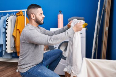 Young latin man smiling confident washing clothes at laundry room