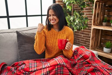 Kanepede oturup kahve içen genç İspanyol kadın çok mutlu ve heyecanlı bir şekilde kollarını kaldırıp, gülümseyerek ve başarı çığlıkları atarak zafer hareketi yapıyor. kutlama konsepti. 