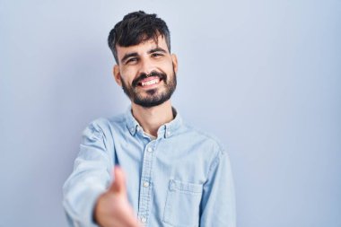 Mavi arka planda duran sakallı genç İspanyol adam selamlama ve hoş geldin demek için dostça el sıkışma teklif ediyor. başarılı iş. 