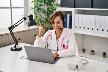 Middle age doctor woman working wearing headset surprised with an idea or question pointing finger with happy face, number one 