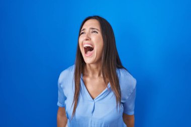 Genç esmer kadın mavi arka planda kızgın ve kızgın bağırıyor sinirli ve öfkeli, öfkeyle bağırıyor. öfke ve agresif konsept. 