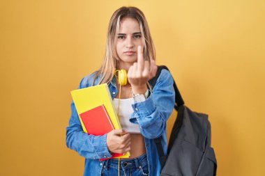 Young blonde woman wearing student backpack and holding books showing middle finger, impolite and rude fuck off expression 