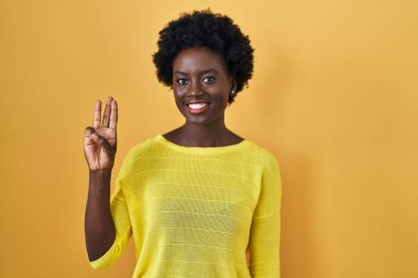 African young woman standing over yellow studio showing and pointing up with fingers number three while smiling confident and happy. 