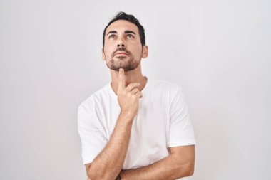 Handsome hispanic man standing over white background thinking concentrated about doubt with finger on chin and looking up wondering 