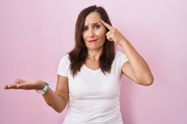 Middle age brunette woman standing over pink background confused and annoyed with open palm showing copy space and pointing finger to forehead. think about it. 