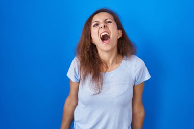 Brunette woman standing over blue background angry and mad screaming frustrated and furious, shouting with anger. rage and aggressive concept. 