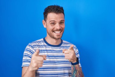 Young hispanic man standing over blue background pointing fingers to camera with happy and funny face. good energy and vibes. 