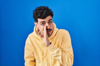 Hispanic man standing over blue background hand on mouth telling secret rumor, whispering malicious talk conversation 
