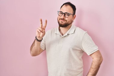 Plus size hispanic man with beard standing over pink background smiling looking to the camera showing fingers doing victory sign. number two. 