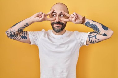 Sarı arka planda dikilen dövmeli genç İspanyol adam barış sembolü yapıyor. Yüzünde parmaklar, gülümseyerek zaferi gösteriyor. 