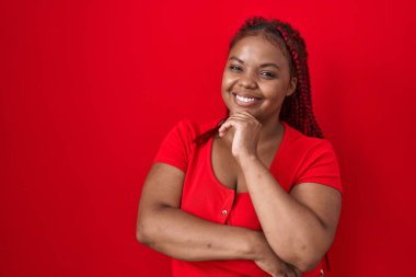 African american woman with braided hair standing over red background looking confident at the camera smiling with crossed arms and hand raised on chin. thinking positive. 